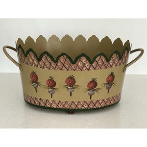 May include: An oval metal planter with a scalloped top edge and two side handles. The exterior is painted a muted gold with a repeating pattern of five red strawberries and a decorative lattice border in pink and dark green along the top and bottom.
