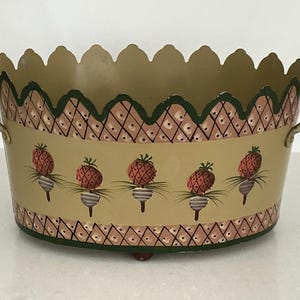 May include: Oval metal container with scalloped rim and handles. The container is painted tan with a repeating pattern of pineapples and a diamond design. The rim is green and the handles are gold.