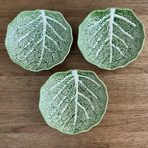 May include: Three green leaf-shaped ceramic dishes. Each dish has a detailed vein pattern in white, resembling a cabbage leaf. The dishes are arranged on a wooden surface, showcasing their naturalistic design. These decorative dishes are ideal for serving small portions.