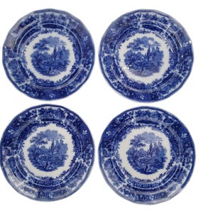 May include: A set of four round ceramic plates featuring a blue and white transferware pattern. Each plate displays a central landscape scene with trees and buildings, surrounded by a decorative floral and scroll border on the scalloped rim.