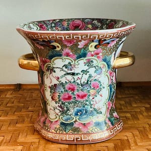 May include: A decorative ceramic vase with a flared top and gold handles. The vase features a colorful floral design with pink, blue, and green flowers, and butterflies. The rim and base have a brown geometric pattern.