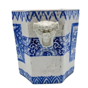 May include: An octagonal ceramic planter featuring a white base with blue decorative patterns and a prominent white relief sculpture of a lion head on the front. The design includes intricate blue floral and geometric borders along the top and bottom.