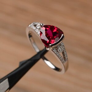 May include: A silver ring with a large, heart-shaped, deep red gemstone as the focal point. The band is adorned with smaller, clear gemstones. The ring is held by a black tool against a blurred wooden background.