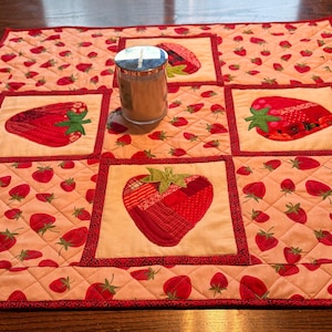May include: A quilted placemat with a repeating strawberry pattern and five large strawberry appliqués. The strawberries are red with green leaves, set against a light pink background. A glass jar sits on the placemat.