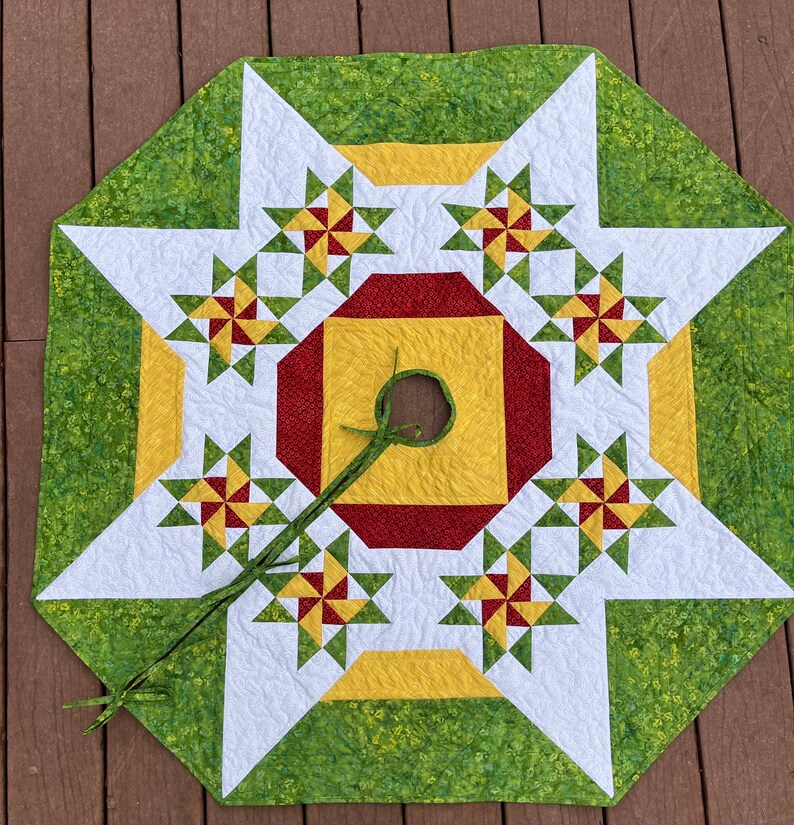 May include: A green, white, red, and yellow quilted Christmas tree skirt with a star design and a hole in the center for a tree stand.