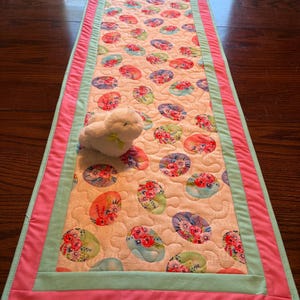May include: A quilted Easter table runner featuring a repeating pattern of floral-filled egg shapes in shades of pink, blue, and green on a cream background. The runner is bordered with a light green and pink fabric.