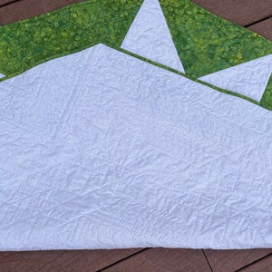 May include: A white quilted fabric with a green and white patterned border. The fabric is folded in half and has a decorative swirl pattern.