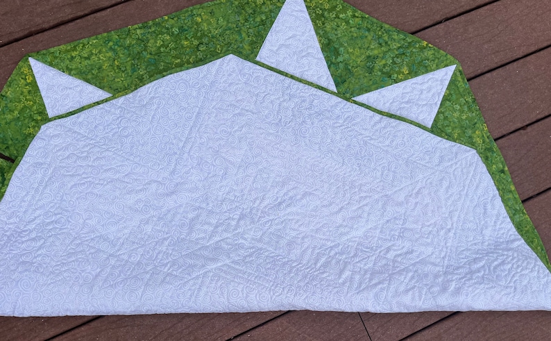 May include: A white quilted fabric with a green and white patterned border. The fabric is folded in half and has a decorative swirl pattern.