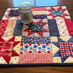 Peut inclure: Un dessous de plat carré, sur le thème patriotique, avec une variété de tissus à motifs rouges, blancs et bleus. Un pot cylindrique avec un couvercle argenté est posé sur le tapis. Les tissus comprennent des étoiles, des rayures et des carreaux.