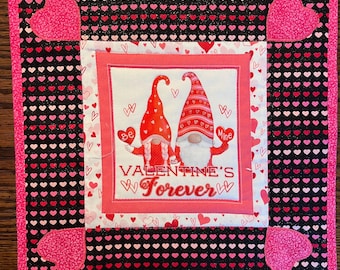 May include: A pink and black quilted square with a heart-shaped border. The center features a white square with a pink border. Inside the white square is a picture of two gnomes with red hats and the text "Be Mine" and "Valentine's Forever".