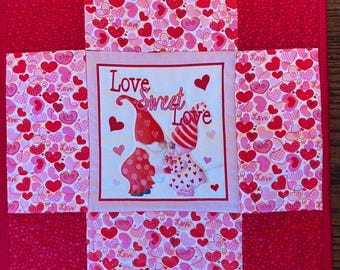 May include: A Valentine's Day themed quilted square featuring a central panel with the words "Love Sweet Love" and illustrations of gnomes. The surrounding fabric is patterned with hearts in shades of pink and red.