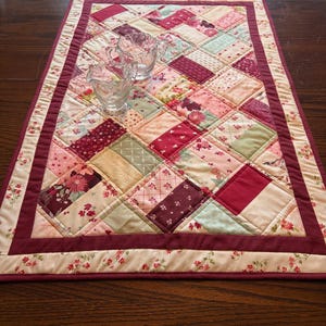 May include: A rectangular quilted table runner featuring a patchwork design of various pink, burgundy, and cream floral and geometric fabric squares, bordered by a solid burgundy frame, with two clear glass sugar bowls placed on top.