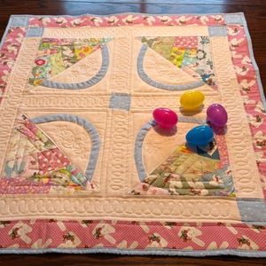 May include: A handmade Easter-themed quilt with a patchwork design. The quilt features pastel colors, floral patterns, and a pink border with bunny prints. Four colorful plastic Easter eggs are placed on the quilt.