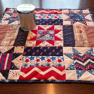 May include: A square patchwork placemat featuring red, white, and blue star and stripe designs. A glass candle sits on the placemat. The placemat has a dark blue border. The candle has a silver lid.