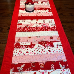 May include: A red and pink striped table runner with a Valentine's Day theme. The runner is made of fabric scraps and features hearts, gnomes, and the words "Love Wins" and "Be Mine".