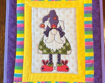 May include: A purple, yellow, green, and pink striped fabric square with a gnome wearing a purple hat and red bunny slippers in the center. The gnome is holding a flower.