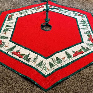 May include: A red Christmas tree skirt with a white border featuring a festive design of red trucks carrying Christmas trees, green trees, and deer. The skirt has a green trim and a tie closure.
