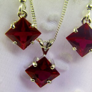 May include: A set of red gemstone jewelry featuring a pendant necklace and earrings. The pendant and earrings each have a square-shaped red gemstone with silver settings.