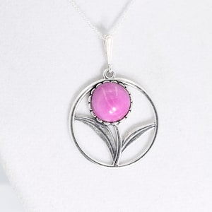 May include: A silver-toned pendant necklace featuring a circular frame with a stylized flower design. The center of the flower is a smooth, round, vibrant pink cabochon stone. The pendant hangs from a delicate silver-toned chain.