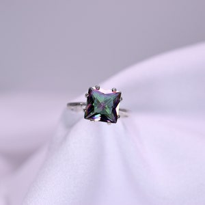 May include: A silver ring with a square-shaped iridescent gemstone. The gemstone has a green, blue, and purple hue.