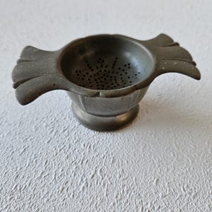 Puede incluir: Un colador de té plateado vintage con un diseño en forma de cuenco y pequeñas perforaciones. El colador tiene dos asas decorativas y una base de pedestal. Este artículo de cocina probablemente está hecho de metal y está diseñado para té de hojas sueltas.