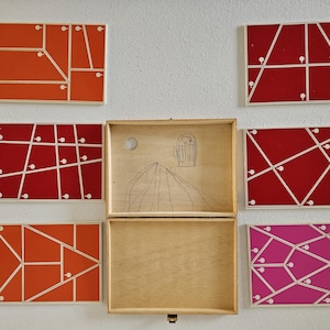 Puede incluir: Una caja de madera con tapa abatible, abierta para revelar un dibujo de una montaña y una casita de pájaros. La caja está rodeada de ocho azulejos coloridos, cada uno con un patrón geométrico diferente y puntos blancos.