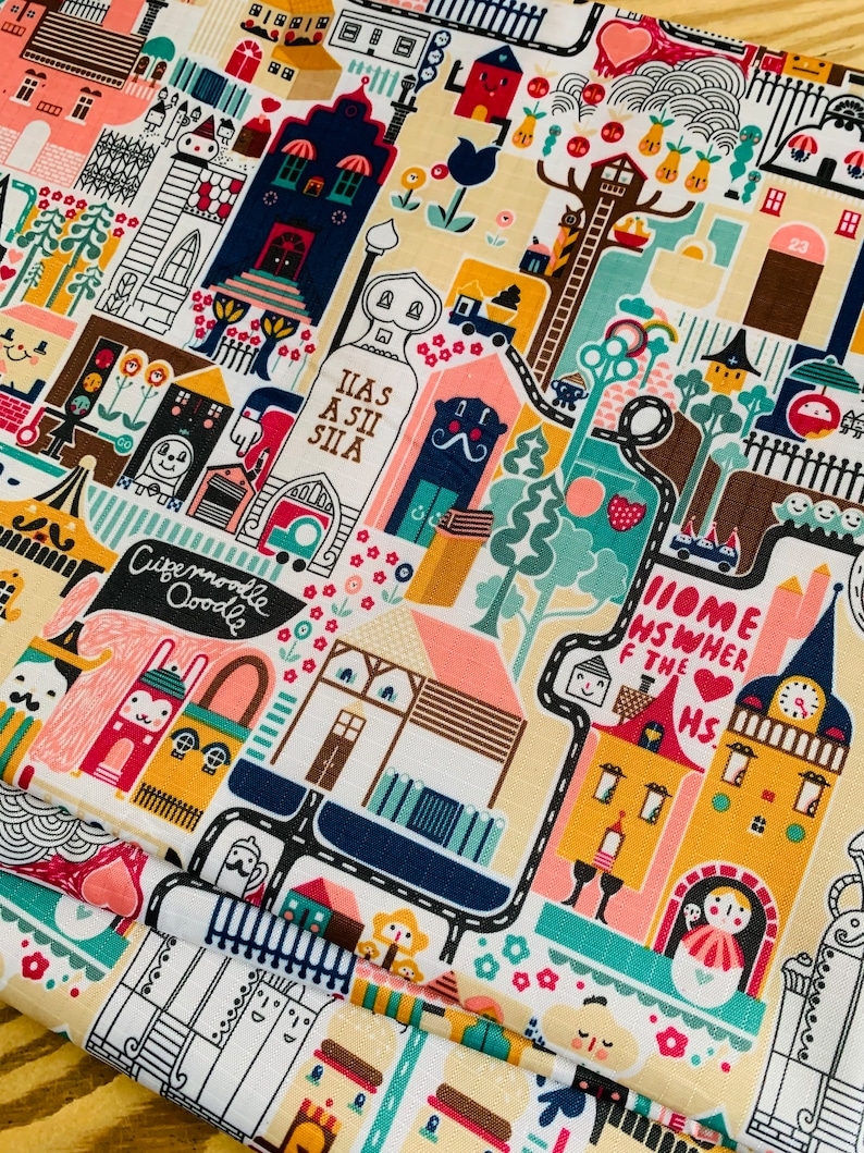 May include: A colorful fabric with a whimsical design featuring houses, trees, and other buildings. The fabric has a white background with black outlines and bright colors. The text "Home is where the heart is" is featured on a yellow building.