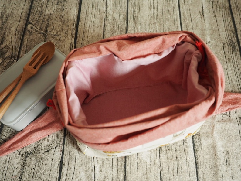 May include: A pink fabric lunch bag with a white lining and a floral pattern. The bag is open and empty.