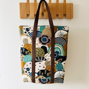 Puede incluir: Una bolsa de lona marrón con un estampado abstracto colorido de colinas, árboles y flores. La bolsa tiene correas marrones y un cierre de cremallera.