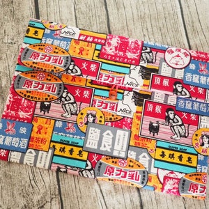 Pode incluir: Uma bolsa de tecido colorida com uma variedade de anúncios de estilo vintage impressos nela. A bolsa apresenta um esquema de cores vermelho, branco e azul com imagens de macacos, fogo e texto em caracteres chineses. A bolsa tem um fecho de botão.