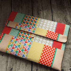Puede incluir: Una funda para portátil de tela de patchwork colorida con cierre de botón. La funda presenta una variedad de patrones, incluyendo rayas, lunares y diseños geométricos en tonos de rojo, amarillo, verde y blanco.