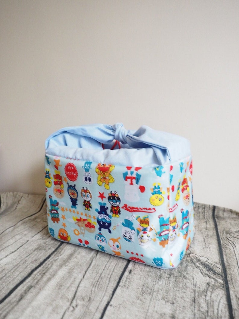 May include: A light blue fabric drawstring bag with a colorful cartoon print featuring characters from the Japanese anime series "Yo-kai Watch".