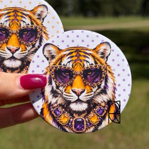 Può includere: Due spille rotonde con l'illustrazione di una tigre. La tigre ha il pelo arancione e nero, indossa occhiali da sole con lenti viola e montatura dorata, e una collana con grandi gemme viola e ambra su uno sfondo bianco a pois viola.