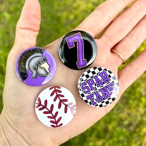 May include: A collection of four round buttons. One button features a purple and white Spartan helmet design. Another has a large purple number 7 on a black background. A third button displays a baseball design with red stitching. The last button says "SPARTANS" on a checkered background.