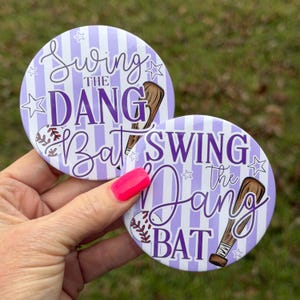 Könnte beinhalten: Zwei runde Baseball-Buttons mit helllila und weiß gestreiftem Hintergrund. Die Buttons zeigen den Text "Swing the Dang Bat" in einer Schreibschrift, zusammen mit einem Baseballschläger und einer Baseball-Grafik.