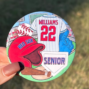 May include: A round button with a baseball theme. It features a red baseball cap with the words "Red Sox" on it, a blue and white baseball jersey with the name "Williams" and the number "22" on it, a blue baseball glove, a baseball bat, and a white home plate with the word "Senior" on it. The background is a green baseball field.