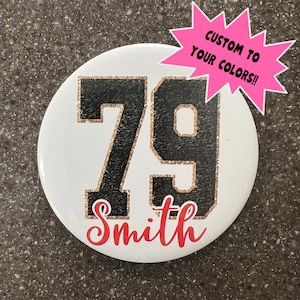 May include: A white button with black glitter numbers 79 and the name Smith in red glitter. The button has a pink starburst sticker with the text "Custom to your colors!!"