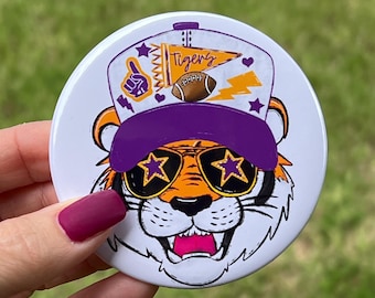 May include: A white round button with a cartoon tiger wearing purple sunglasses with gold stars, a purple and white baseball cap with the word "Tigers" on it, and a football.
