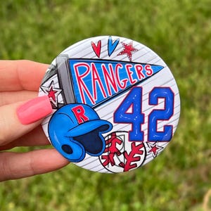 May include: A circular button featuring a blue pennant with the text RANGERS in red, a blue baseball helmet with a red R, the number 42 in blue, and red star and baseball graphics on a white background with thin diagonal gray lines.