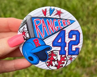 May include: A circular button featuring a blue pennant with the text RANGERS in red, a blue baseball helmet with a red R, the number 42 in blue, and red star and baseball graphics on a white background with thin diagonal gray lines.