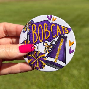 May include: A round button featuring a purple pennant with the yellow text BOBCATS, a purple and yellow megaphone with the name REESE in yellow, purple and yellow pom-poms, and small purple and orange heart icons on a white background.