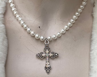 Op de afbeelding: Een parelketting met een zilveren kruisvormige hanger. Het kruis is versierd met kleine, iriserende kristallen en ingewikkelde details. De ketting wordt getoond op een mannequin met een witte, harige rand.