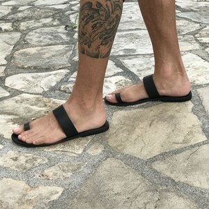 May include: A pair of black leather sandals with a toe thong and a single strap across the foot. The sandals are worn on a person's feet, standing on a stone surface.
