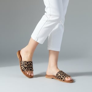 May include: A pair of flat slide sandals featuring a brown and black leopard print pattern on the wide top strap, paired with white cropped trousers. The sandals have a simple open-toe design with a flat brown sole and a textured animal print finish.