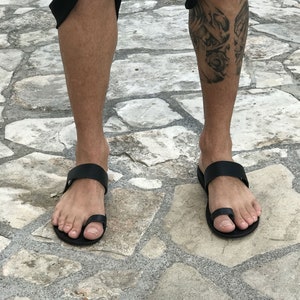 May include: A pair of black leather thong sandals with a toe loop. The sandals are worn on a person's feet, standing on a stone surface.