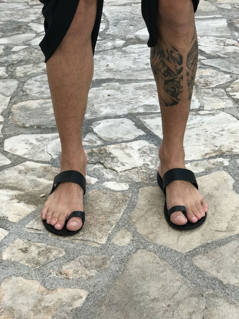 May include: A pair of black leather thong sandals with a toe loop. The sandals are worn on a person's feet, standing on a stone surface.