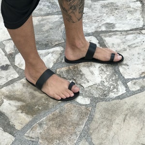 May include: A pair of black leather sandals with a toe loop and a strap across the foot. The sandals are worn on a person's feet, standing on a stone surface.