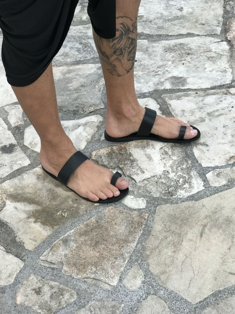 May include: A pair of black leather sandals with a toe loop and a strap across the foot. The sandals are worn on a person's feet, standing on a stone surface.