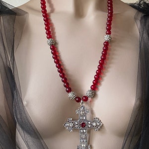 Può includere: Una collana di perline di vetro rosso con accenti argentati e un grande ciondolo a croce d'argento con un gioiello rosso al centro.