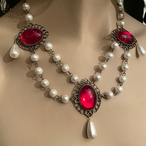 May include: A double strand pearl necklace with three ornate, gold-toned pendants featuring large, red, oval-shaped gemstones. Each pendant has a single pearl drop.